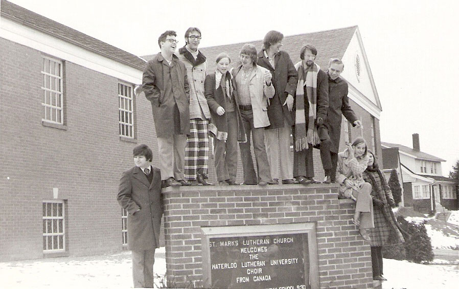 Eight young people stand on top of a church sign. Three young people stand next to it. They are laughing and smiling.